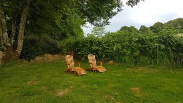 To træstole i Adirondack-stil i et græsfuldt udendørsområde med træer og buske i baggrunden.