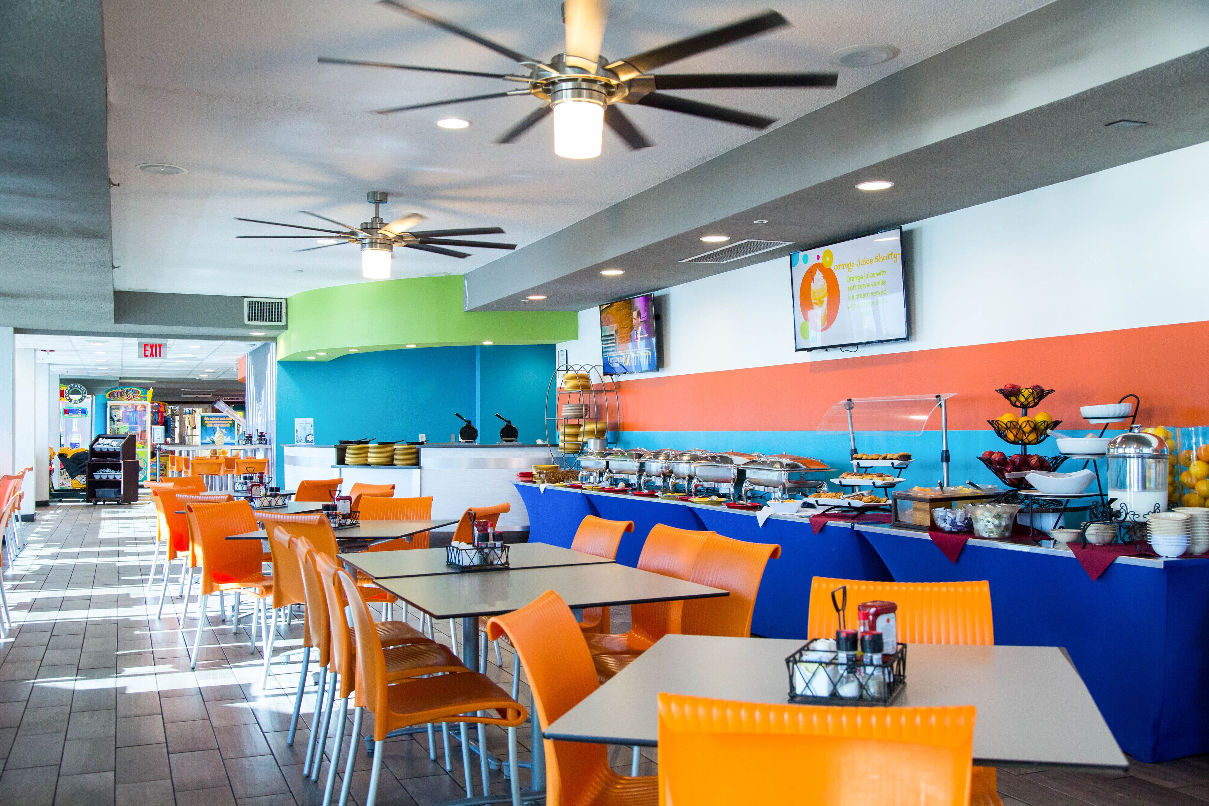 A brightly lit cafeteria with orange and blue seating, a buffet counter with various food items, and ceiling fans.