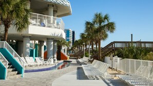 A resort with a pool, lounge chairs, palm trees, and a staircase leading to a balcony.