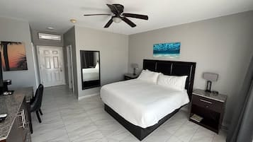 Quarto de hotel com cama, escrivaninha, cadeira, mesa de cabeceira, abajur, ventilador de teto e um quadro na parede.