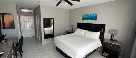 A hotel room with a bed, a desk, a chair, a nightstand, a lamp, a ceiling fan, and a painting on the wall.