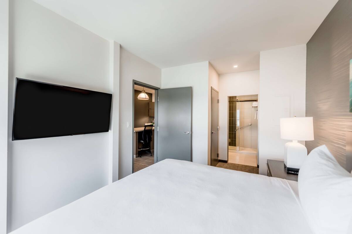 A modern hotel room with a large bed, a flat-screen TV mounted on the wall, a desk with a chair, and a bathroom visible through an open door.