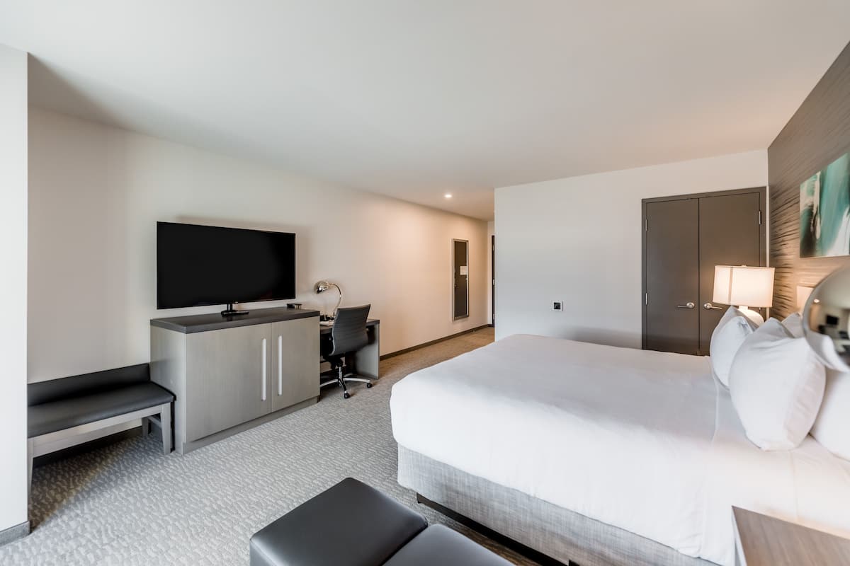 A modern hotel room with a large bed, a desk with a chair, a television, and a wardrobe.
