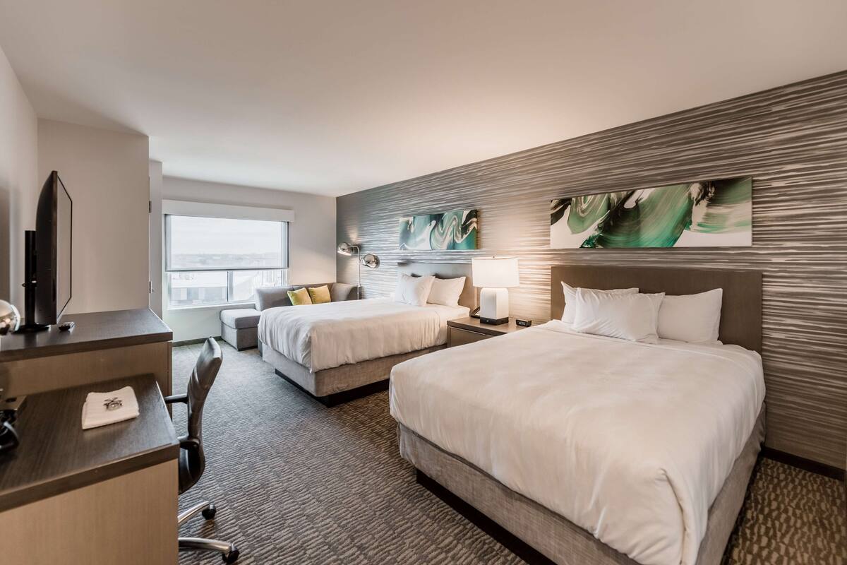 A hotel room with two beds, a desk, a chair, a TV, and artwork on the walls.