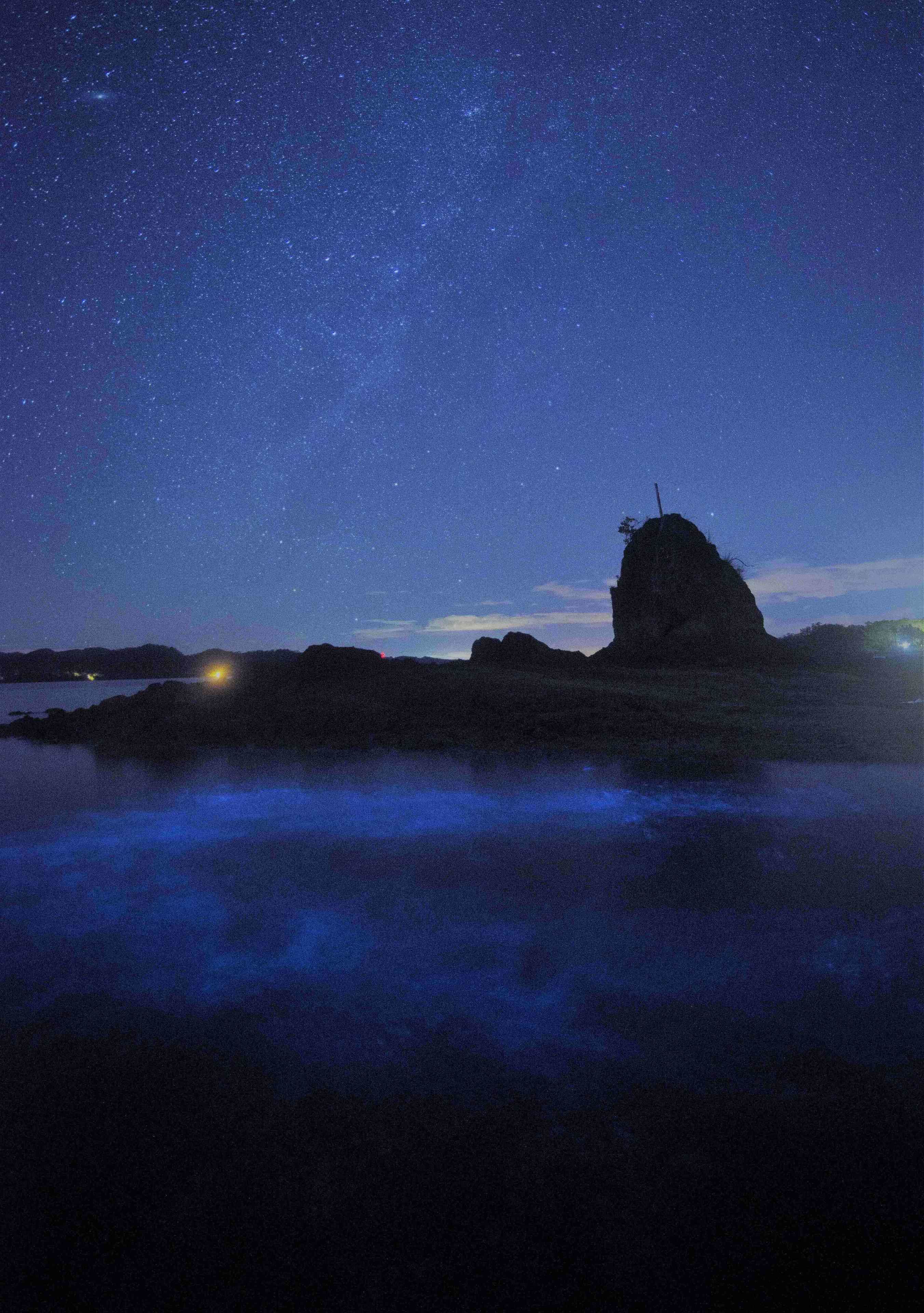 A night scene with a starry sky, a prominent rock formation, and a body of water reflecting the sky.