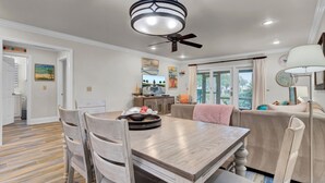A dining area with a wooden table, chairs, and a ceiling fan. There is a kitchen area with cabinets and a window. - 6C Mariners Walk | AvantStay | Easy Pool + Beach Access (Isle of Palms)