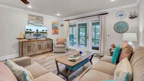 A spacious living room with a wooden coffee table, beige sofas, a wooden cabinet, and a ceiling fan. - 6C Mariners Walk | AvantStay | Easy Pool + Beach Access (Isle of Palms)
