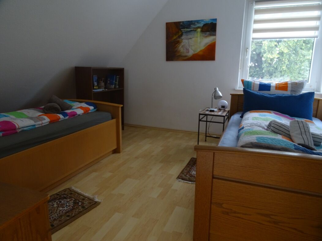 Un dormitorio con dos camas, una mesita de noche con lámpara, una estantería y una ventana con persianas.