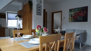 Zona pranzo con tavolo in legno apparecchiato per quattro, circondato da sedie in legno. Si nota un orologio a parete, un quadro incorniciato e un dipinto di fiori.