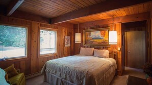 A wooden cabin bedroom with a large bed, two windows, a painting, and a door. - McCall Lakefront Cabin with Dock & Stunning Views (McCall)