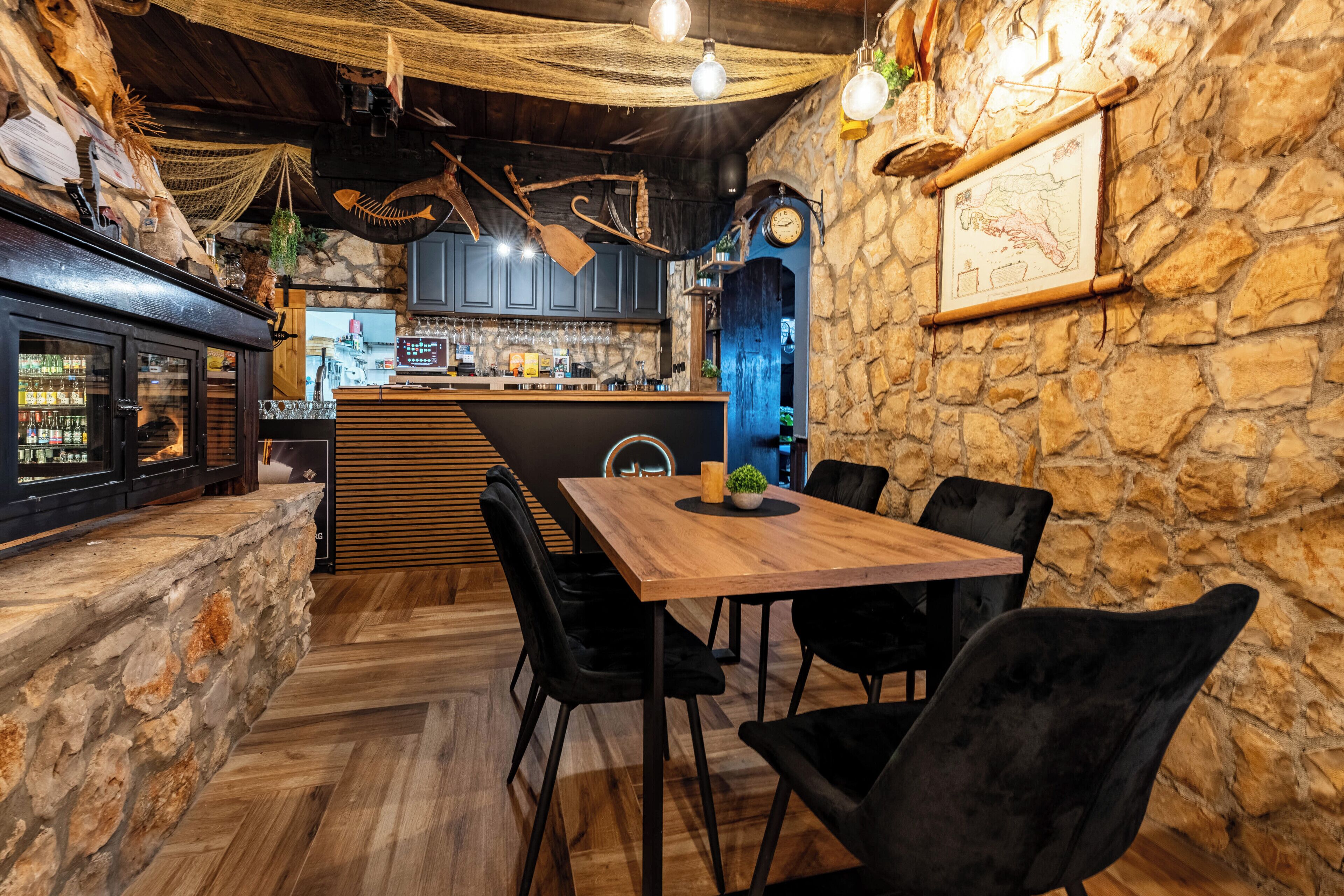 Ein rustikales Interieur mit Holztisch, schwarzen Stühlen, einer Steinwand und einer Bar mit einer Uhr und einer Speisekarte.