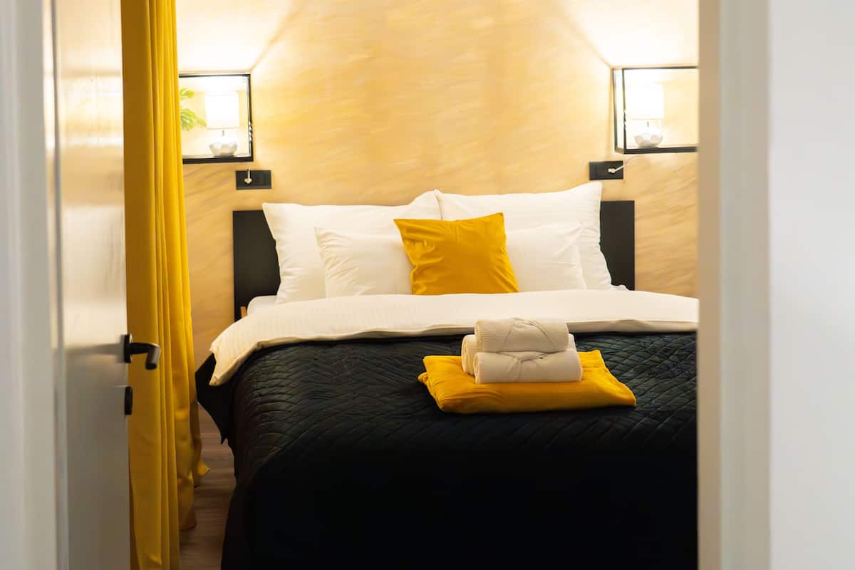 Quarto de hotel com cama, cortina amarela e mesa de cabeceira com abajur.