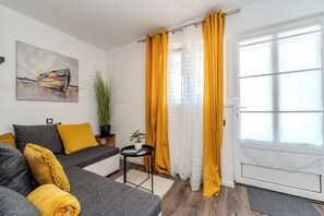 Una sala moderna con un sofá gris, cojines amarillos, un cuadro, una planta en maceta y un ventanal con cortinas amarillas.