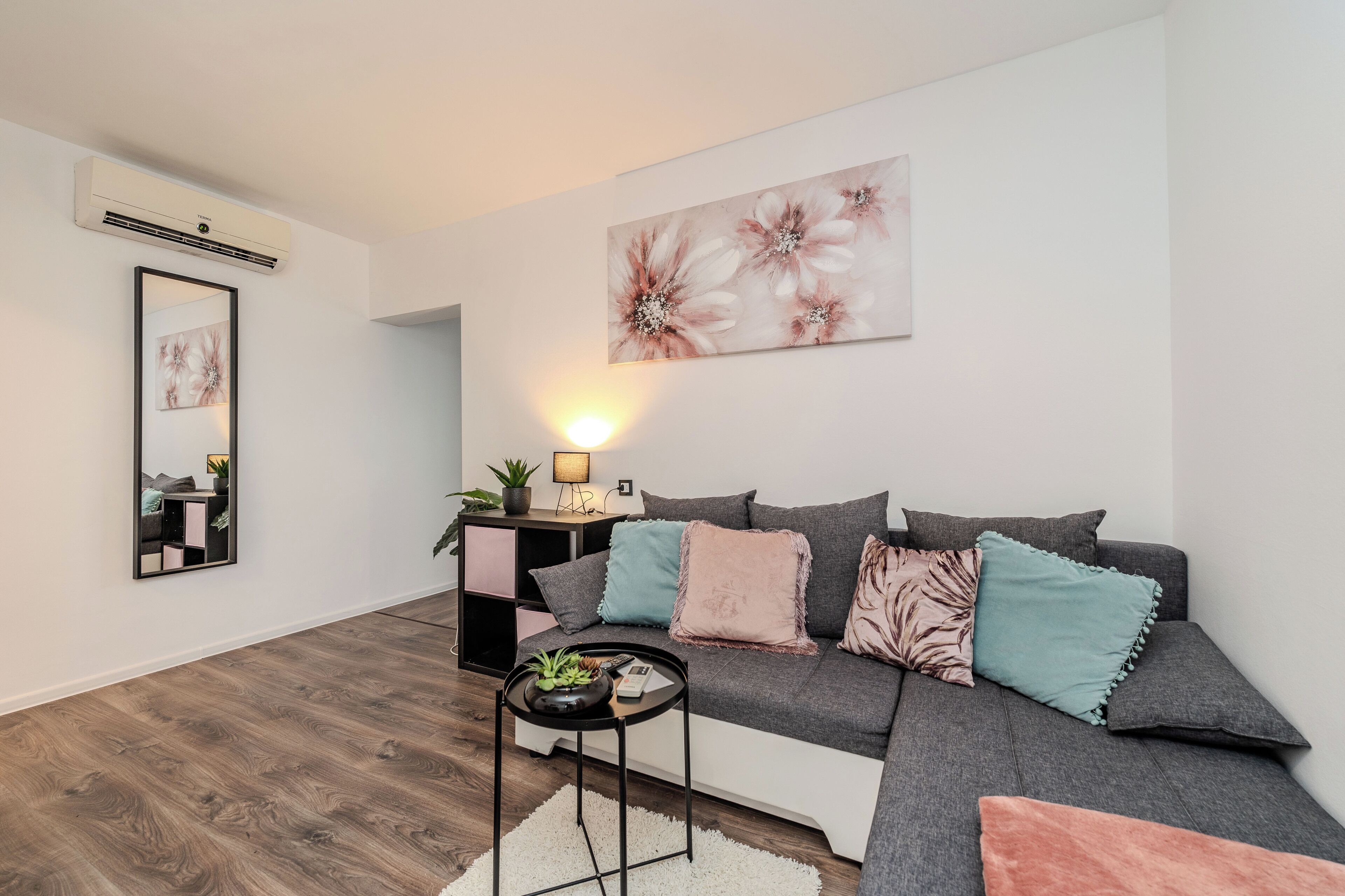 Een moderne woonkamer met een hoekbank, een bijzettafeltje, een vloerlamp en een groot bloemenkunstwerk aan de muur.