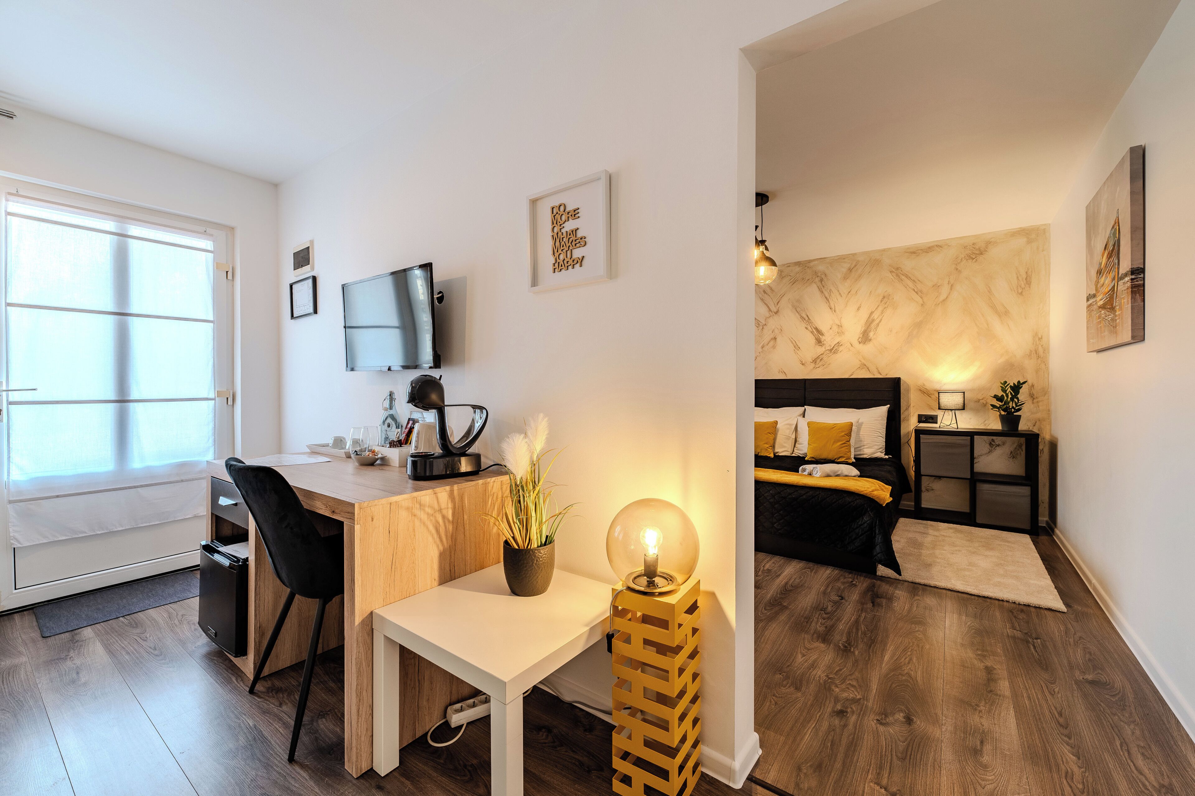 A modern apartment with a wooden desk, a small white table, a bed with a black headboard, and a large window.