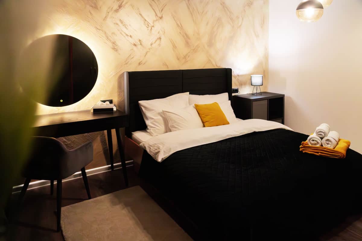 Quarto de hotel com cama, escrivaninha, cadeira, mesa de cabeceira com abajur e espelho redondo.