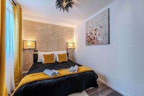 Un dormitorio moderno con colcha negra, dos almohadas blancas y una almohada amarilla de acento. Hay dos lámparas de noche, un cuadro abstracto grande en la pared y una ventana con cortinas.