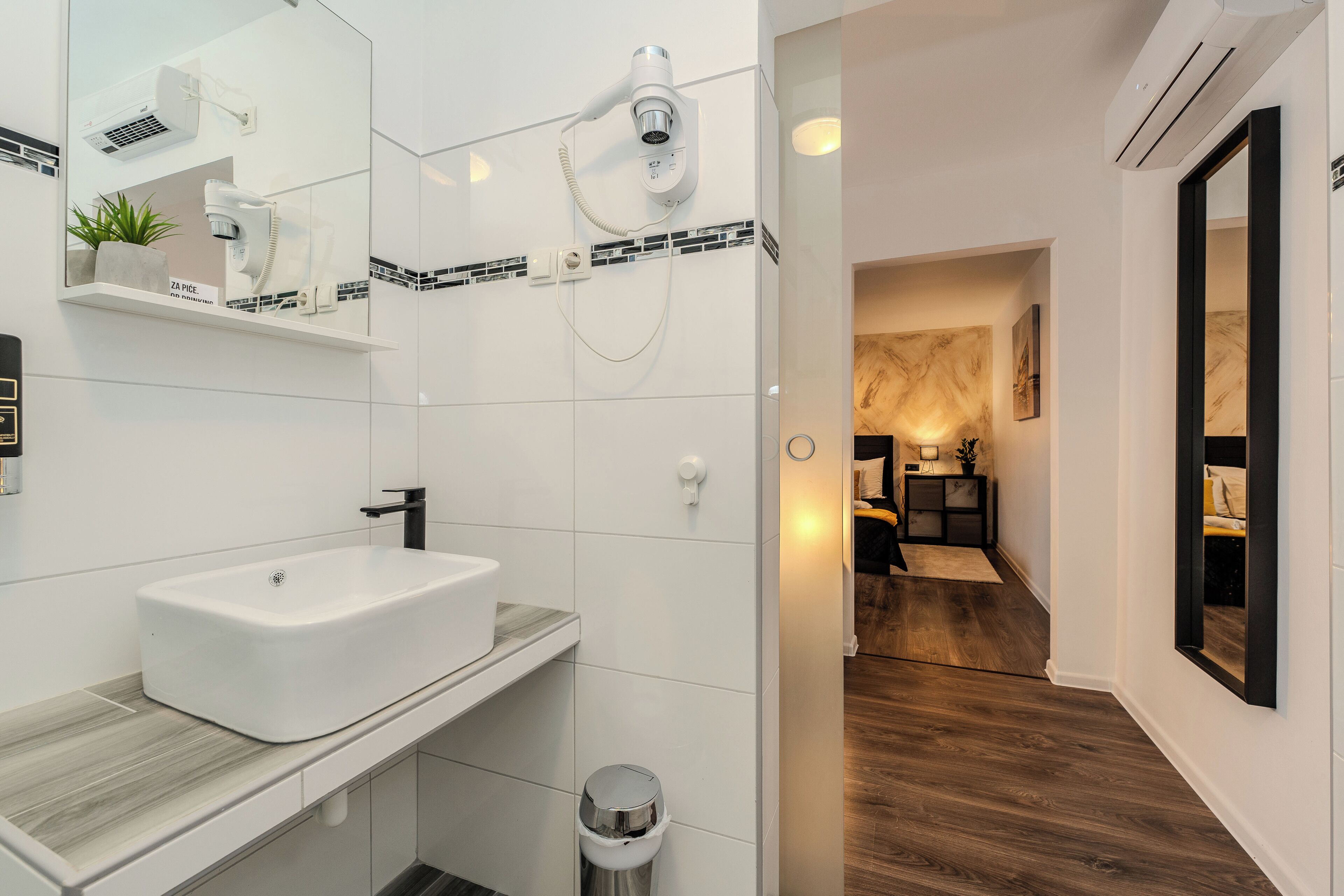 Egy modern fürőszoba fehér mosdókagylóval, tükörrel és törölközőtartóval. A fürőszobában zuhanyzó található kézi zuhanyfejjel, valamint egy WC. A fürőszoba padlóját fa borítja, és a mosdó fölött egy kis növény áll.