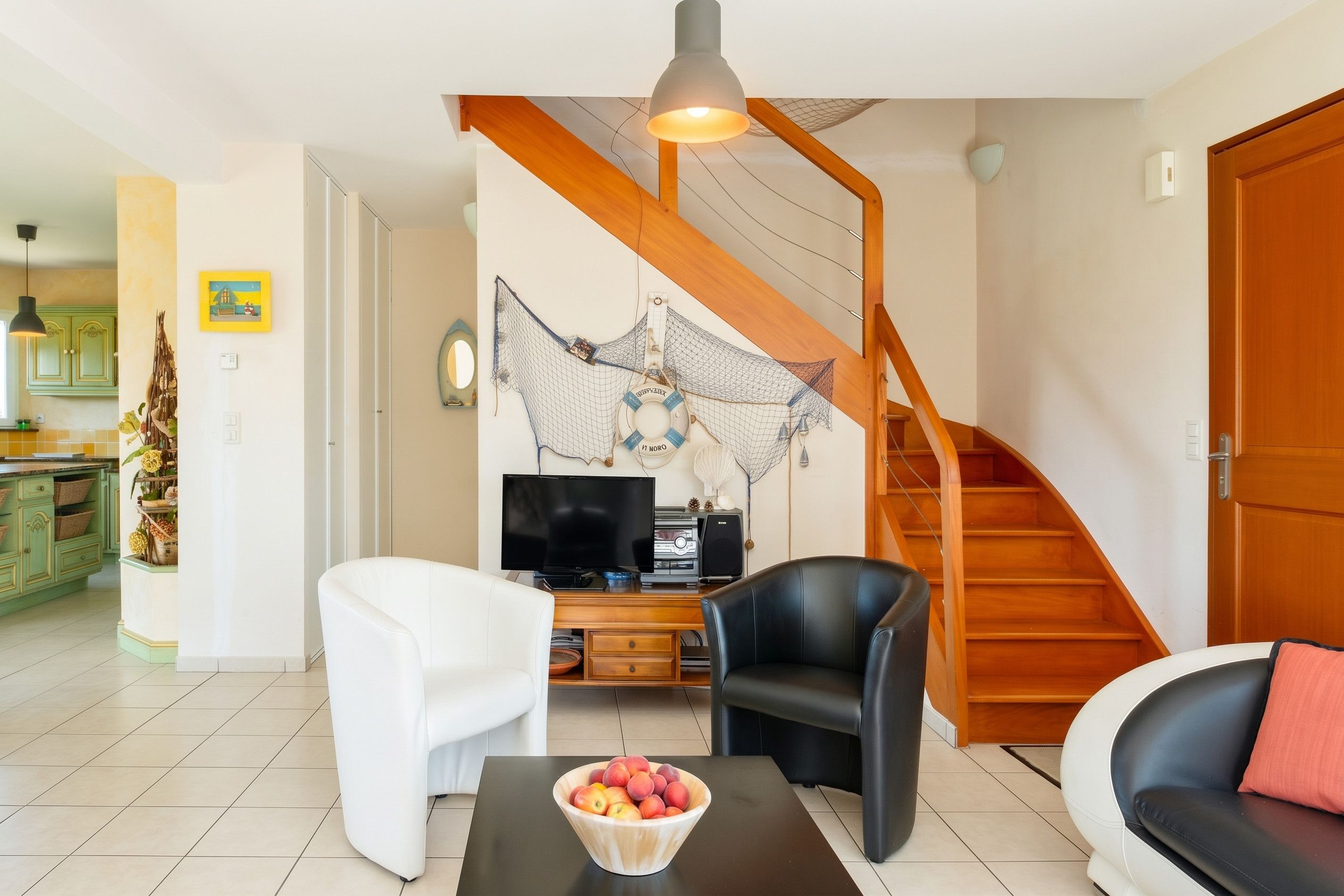 A living room with a TV, two chairs, a coffee table with fruit, a wooden staircase, and a kitchen in the background.