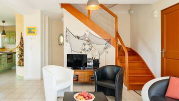 A living room with a TV, two chairs, a coffee table with fruit, a wooden staircase, and a kitchen in the background.