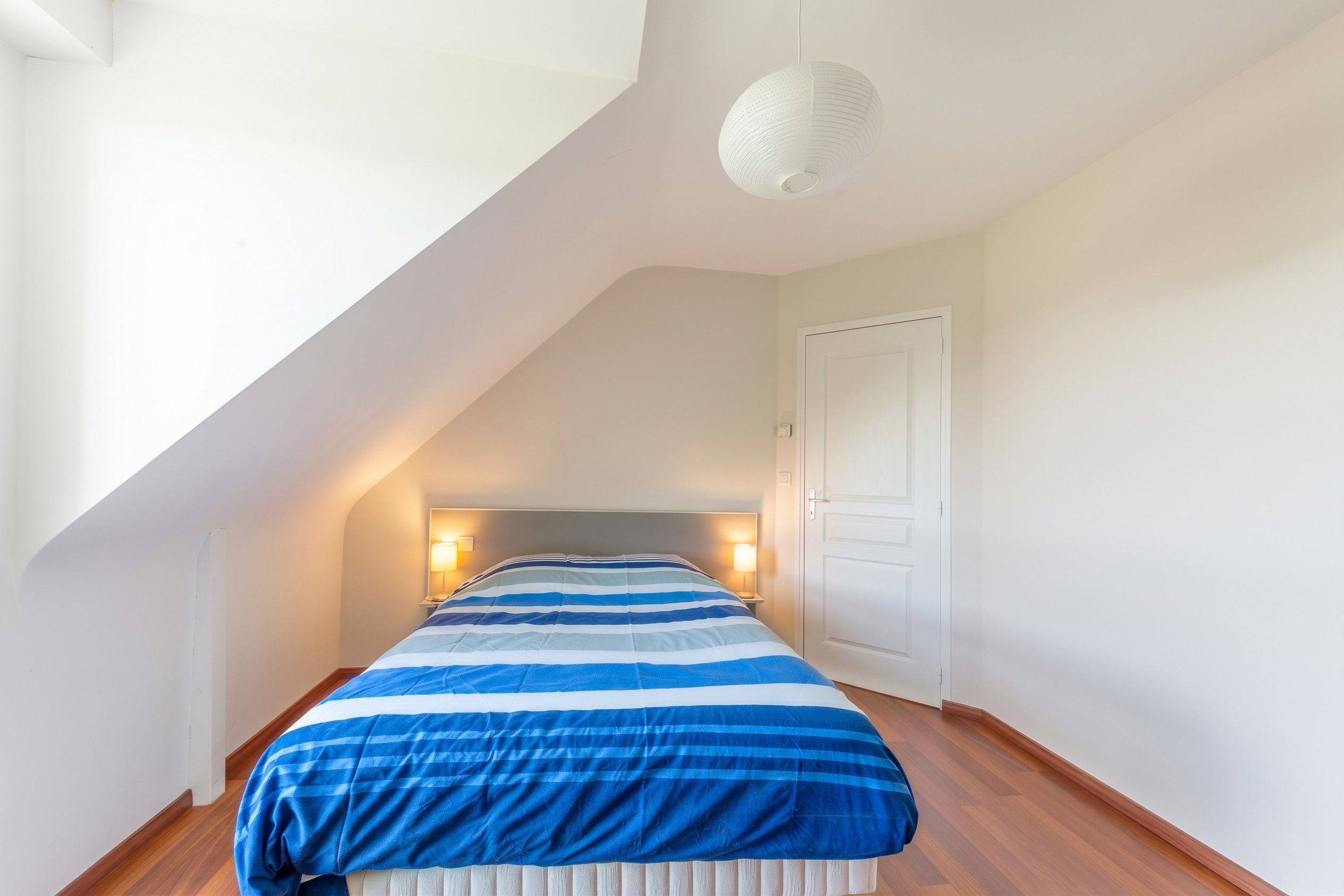 Ein Schlafzimmer mit geneigter Decke, einem Bett mit gestreifter Bettdecke, Nachttischlampen und einer weißen Tür.