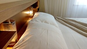Uma cama bem arrumada com lençóis brancos, cabeceira de madeira e mesa de cabeceira com abajur.
