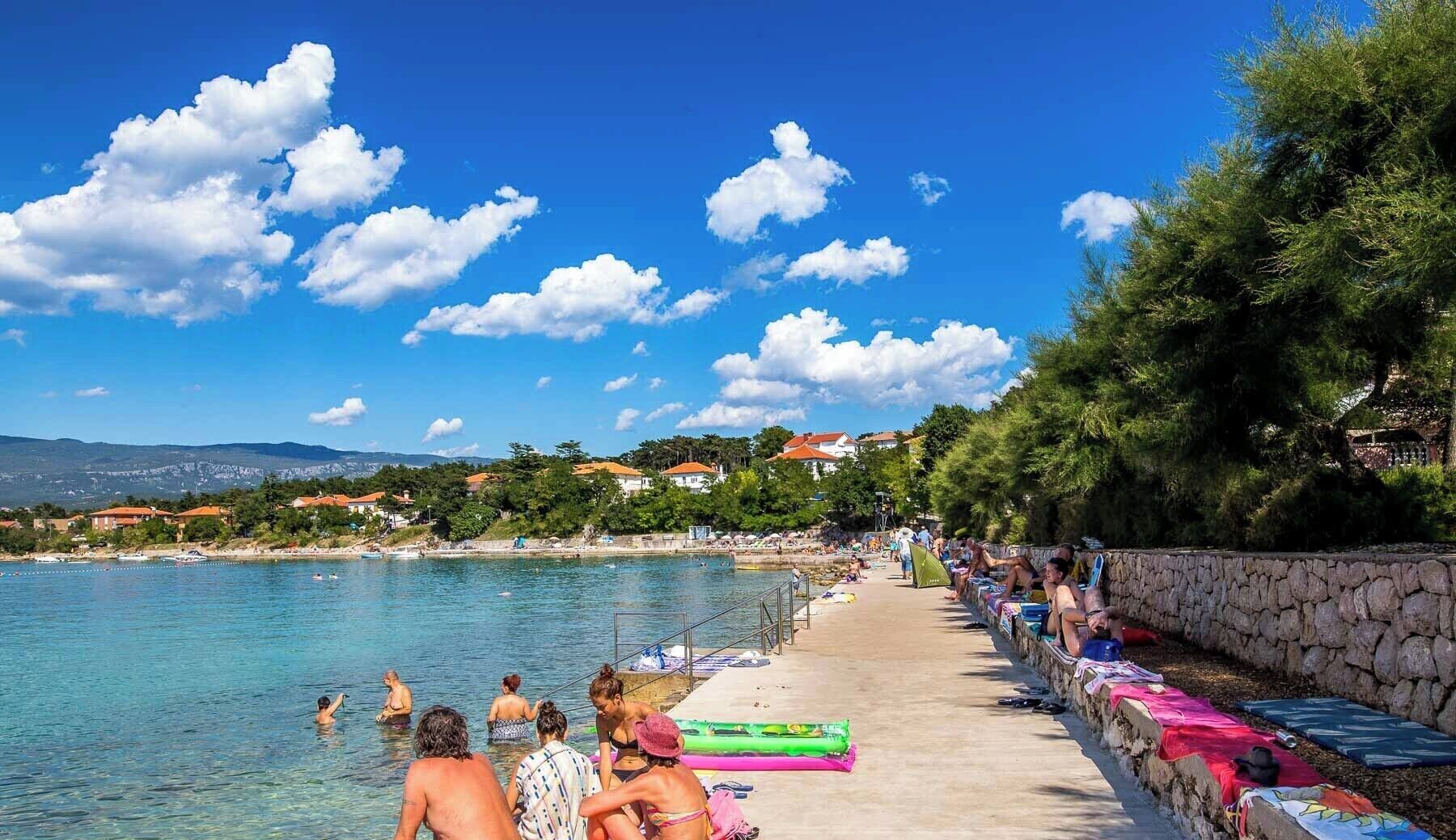 A beach with clear blue water, people swimming and sunbathing, and houses in the background.