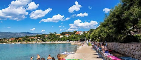 A beach with clear blue water, people swimming and sunbathing, and houses in the background.