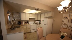 Une cuisine compacte avec des meubles blancs, un four à micro-ondes, une cuisinière et un réfrigérateur. On y trouve une table à manger avec des chaises et un vase de fleurs.