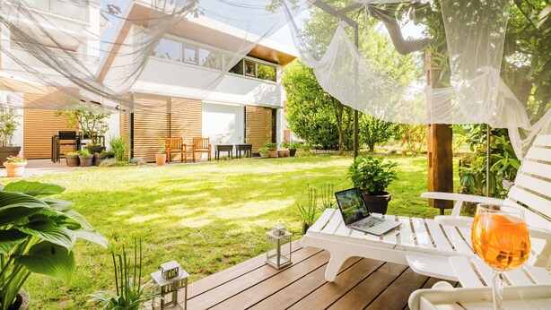 A modern outdoor patio with a wooden deck, white lounge chairs, a laptop, and a glass of wine.