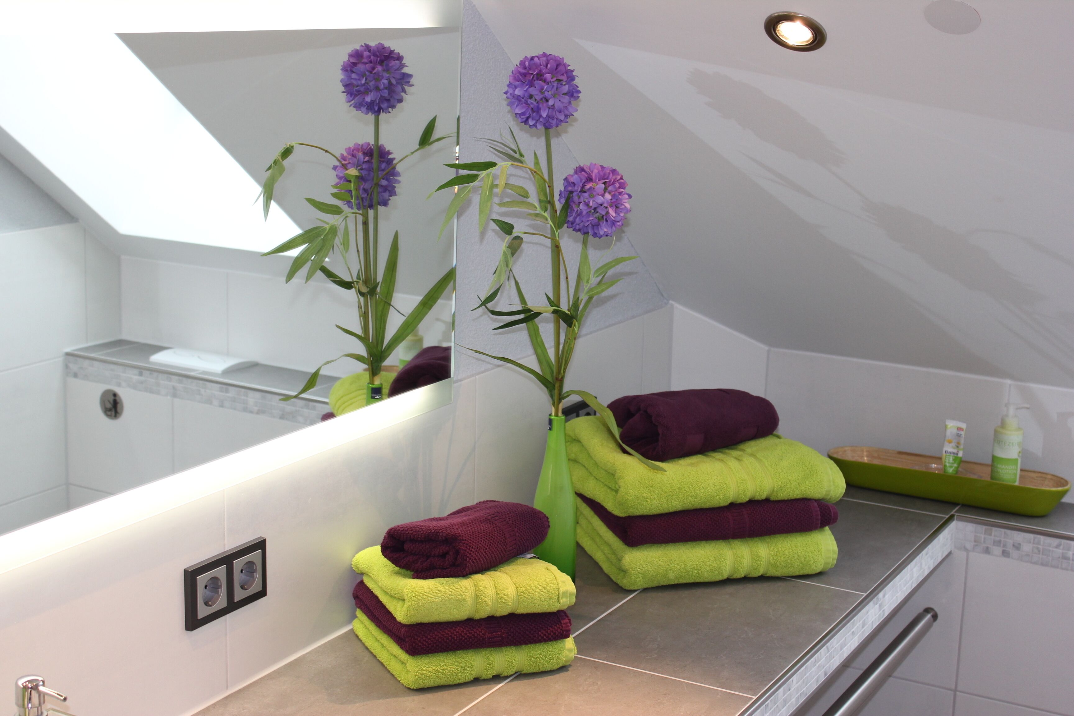 A bathroom with a mirror, a green vase with purple flowers, and stacked towels.