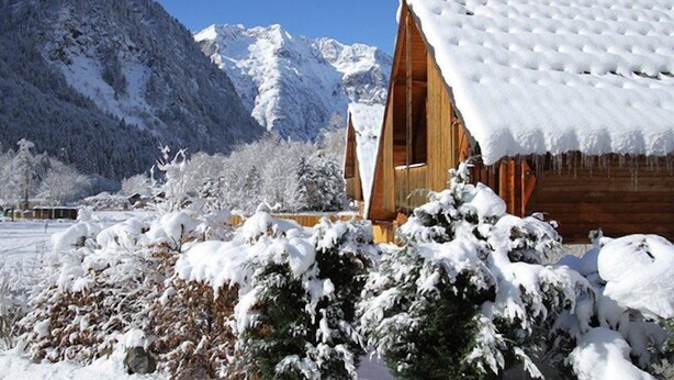 Eine Holzhütte mit geneigtem Dach, umgeben von schneebedeckten Bäumen und Bergen im Hintergrund.