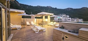 A rooftop terrace with sun loungers, umbrellas, a small table, and a kitchen area.