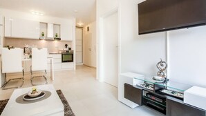 A modern kitchen with white cabinetry, a dining area with white chairs, and a flat-screen TV mounted on the wall.