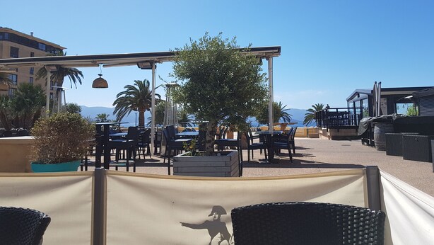 Un espace extérieur aménagé avec des tables et des chaises, une pergola décorée de guirlandes lumineuses et une vue sur la mer.