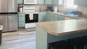 A kitchen with light blue cabinets, a stainless steel refrigerator, a microwave, an oven, and a sink. There is a wooden floor and a large island with stools. - Lakefront Retreat Sleeps 19 • Hot Tub + Paddle Boards • Family & Group Favorite! (Eldon)