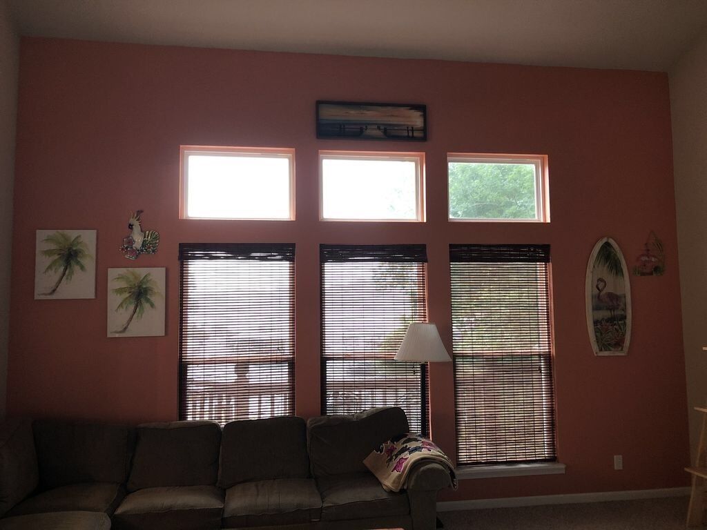 Ein Wohnzimmer mit einer Couch, drei Fenstern mit Jalousien und Wandbildern, darunter ein Hahn und Palmen.