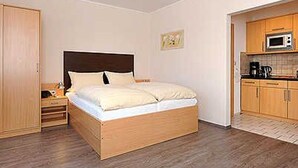 A bedroom with a wooden bed, a nightstand, a wardrobe, and a kitchenette with a microwave. - Apartment 16 - Castle Restaurant (Werdum)
