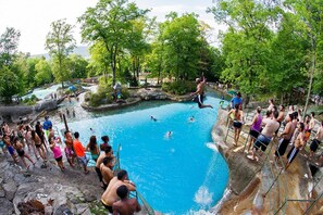 Une piscine naturelle entourée d’arbres et d’un paysage rocheux, où l’on peut voir des gens nager et se tenir debout.