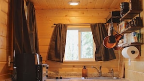 A compact kitchen with wooden walls, a window with curtains, a sink, a coffee maker, and shelves with jars and containers. - A tiny cabin with big adventures (Forks)