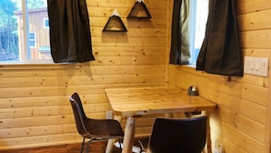 A wooden cabin interior with a wooden table and two chairs, a window with curtains, and a wall-mounted light fixture. - A tiny cabin with big adventures (Forks)