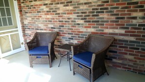 Deux fauteuils en osier avec des coussins bleus et une petite table, placés contre un mur de briques dans un espace extérieur abrité.
