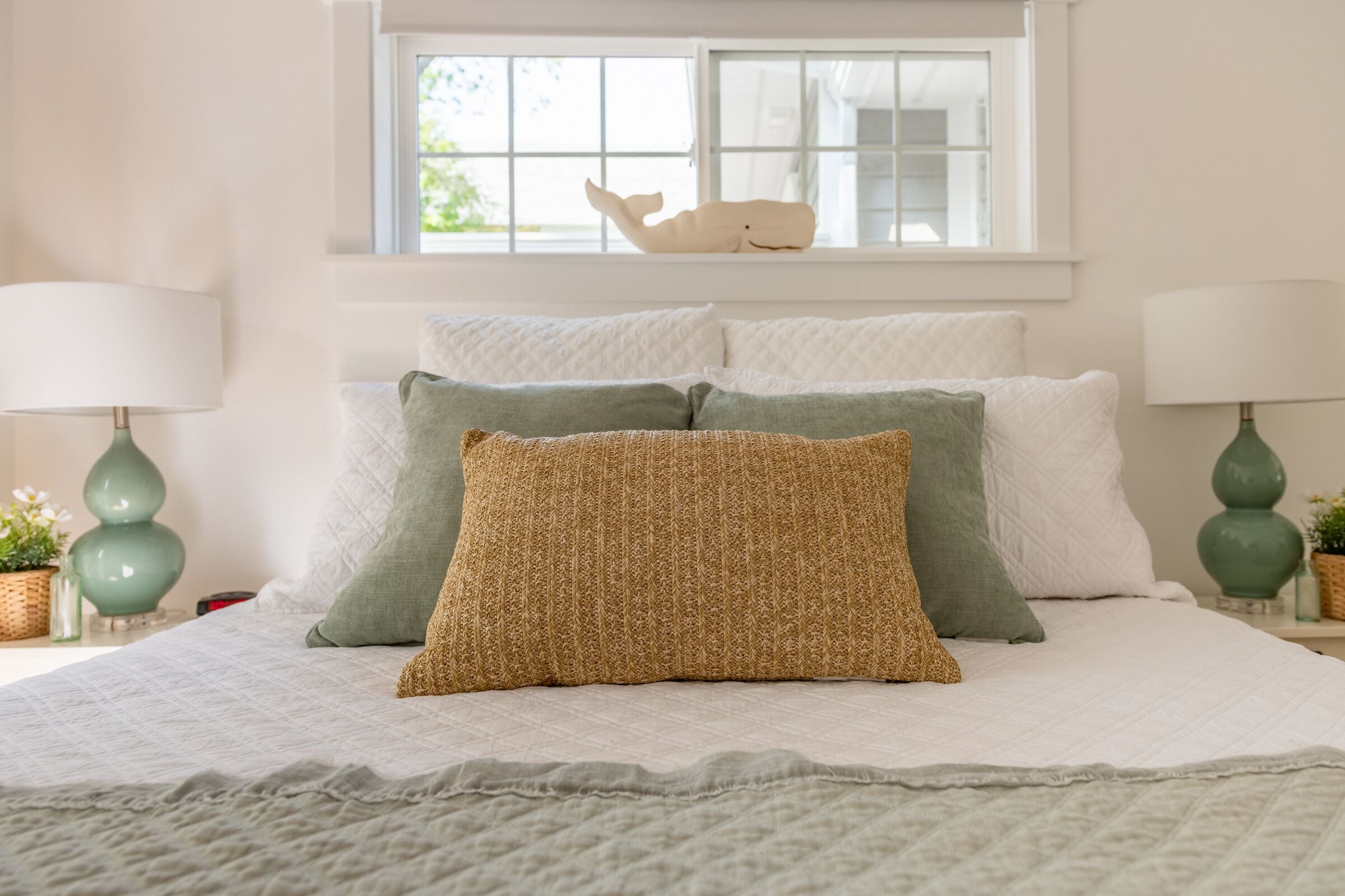 Una cama bien tendida con colcha blanca, almohadas verdes y beige, y una ventana con un adorno decorativo de ballena.