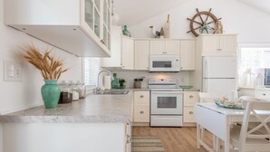 Une cuisine moderne avec des meubles blancs, un réfrigérateur blanc, un four à micro-ondes et une cuisinière. Un décor mural représentant un gouvernail de bateau en bois orne le mur.