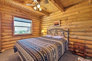 Dormitorio tipo cabaña de madera con cama, mesita de noche, lámpara, ventilador de techo y ventana con vistas.