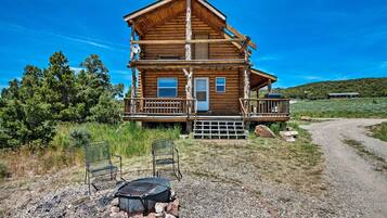 A wooden cabin with a balcony, a fire pit, and two chairs in front.