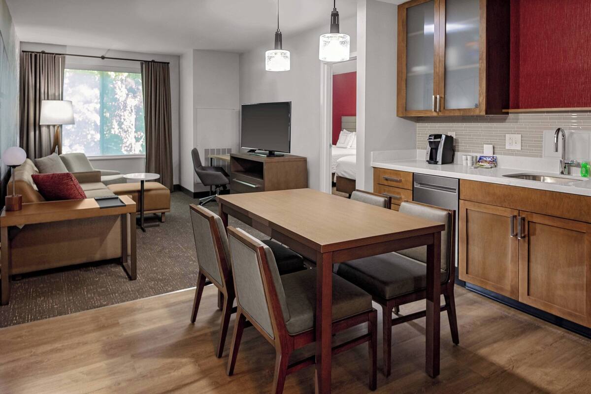 Habitación de hotel con comedor, televisor, sofá y cocina con armarios de madera.