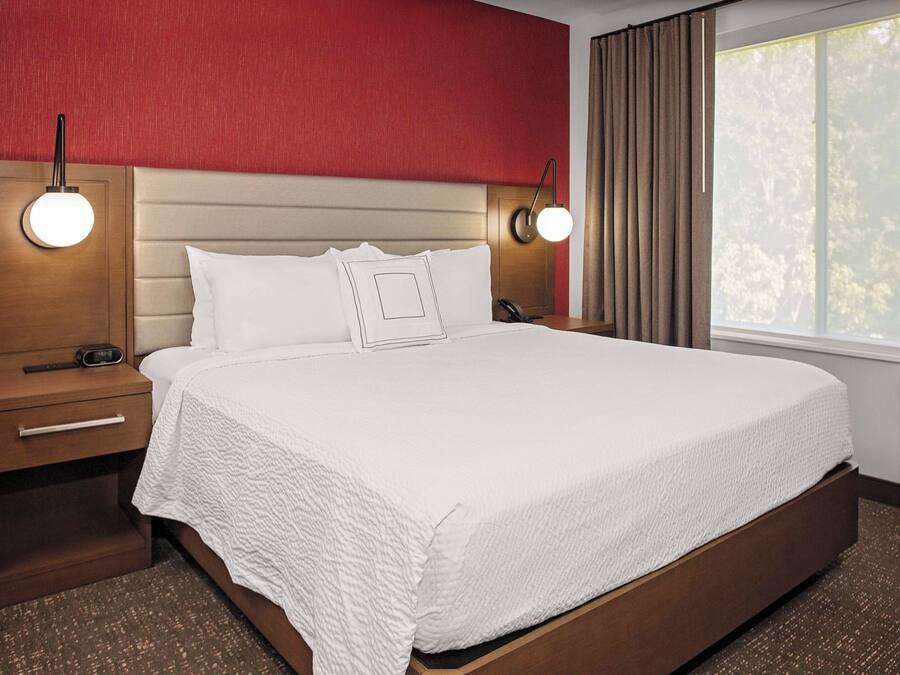 Une chambre d’hôtel avec un grand lit, une table de chevet en bois, une lampe de chevet et une fenêtre avec des rideaux.