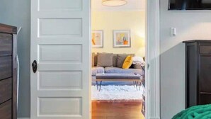 A room with a white door, a grey sofa with cushions, a white rug, and a wooden bench. - Walkable Urban Retreat - 2 En-Suite Baths - Near Top Dining & Shops & Hotspots! (SF)