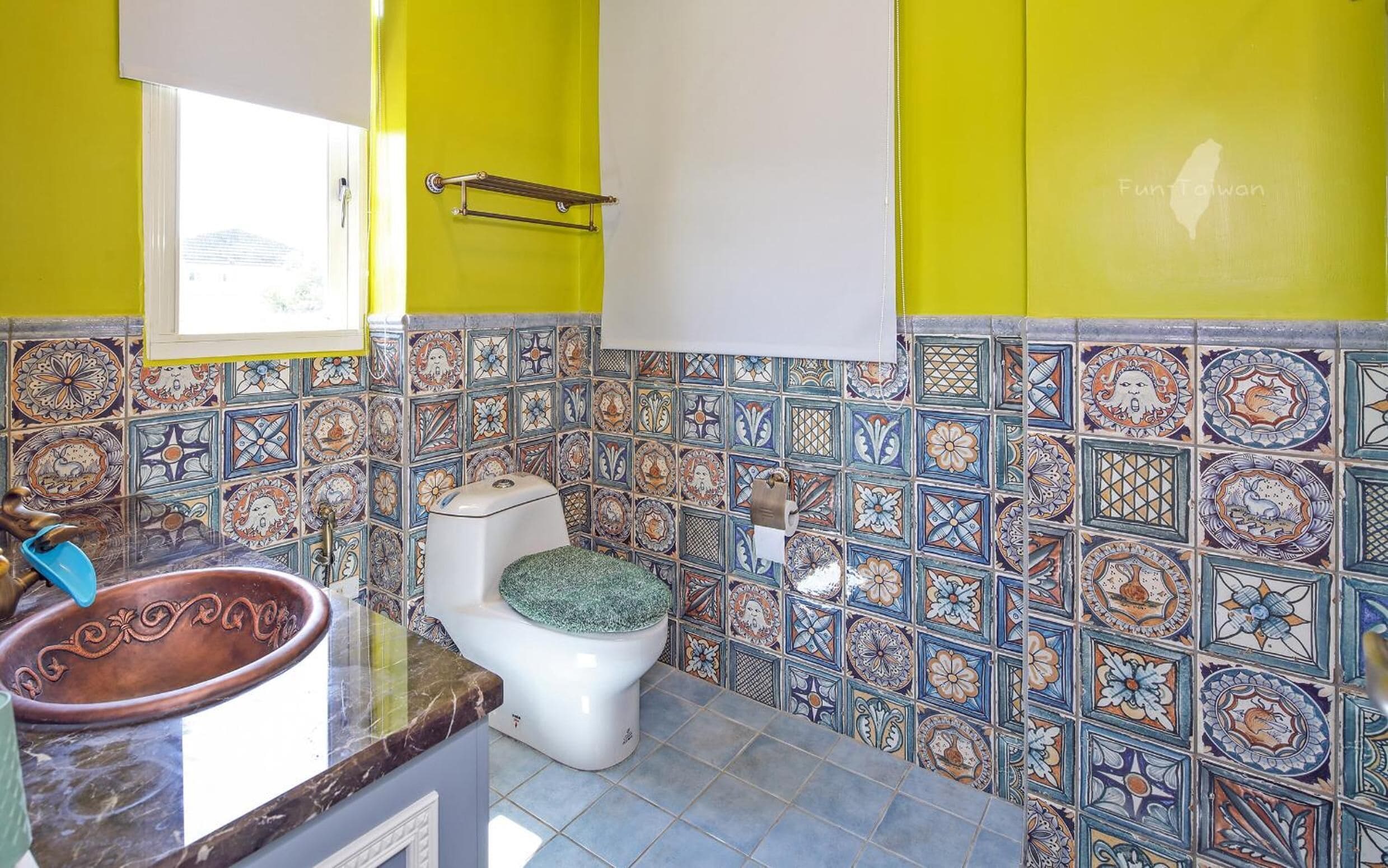 A bathroom with a toilet, a sink, and a window. The walls are tiled with colorful patterns.